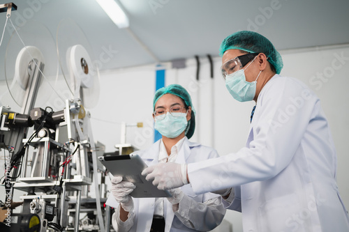 Both male and female Asian scientists are outfitted with protective gear. Examining the factory's machinery and materials used in the mask-making process. Covid 19 is a piece of preventive equipment.