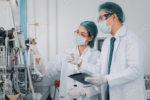 Both male and female Asian scientists are outfitted with protective gear. Examining the factory's machinery and materials used in the mask-making process. Covid 19 is a piece of preventive equipment.