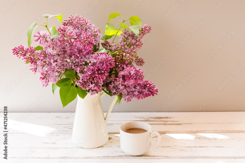 fresh branches of lilacs in a jug on a white wooden table with a cup of shaded coffee. fresh branches of lilacs in a jug on a white wooden table with a cup of shaded coffee.