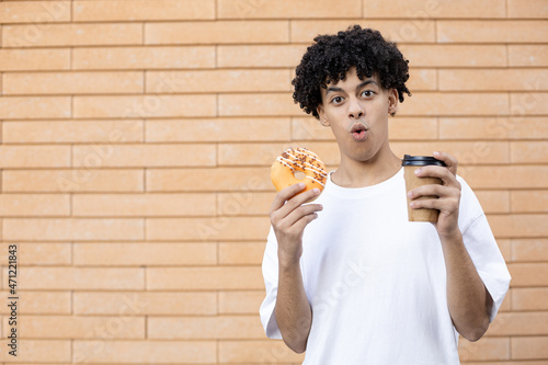 Dessert, drinks, leisure and people concept - Surprised American guy holding a cup of coffee and an orange glazed donut, looking at the camera and wearing a white T-shirt on a brick wall