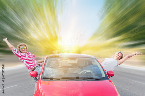 Two wild and crazy Caucasian women with their rental car on the freeway sitting in the doors sticking their bodies out of the vehicle. Thelma and Louise type wild vacation concept