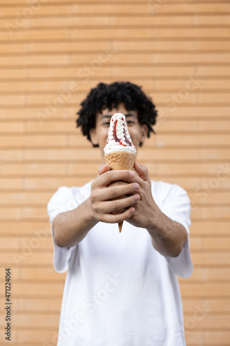 Dessert, junk food and people concept - Close-up of a young curly-haired African-American man holding a white ice cream on his outstretched arms, wearing a white T-shirt on a brick wall background