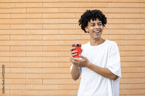 Drinks, lifestyle, consumption and people concept - Laughing curly-haired African-American guy holding a red cup of coffee, wearing a white T-shirt and looking away on a brick wall background