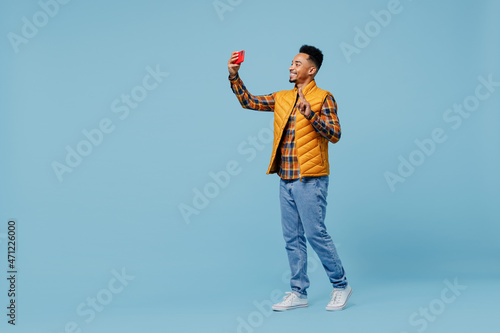 Full size body length young black man 20s wears yellow waistcoat shirt do selfie shot on mobile cell phone post photo on social network isolated on plain pastel light blue background studio portrait.