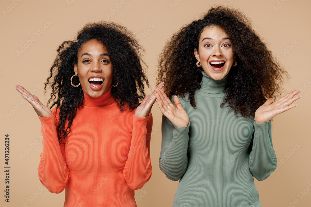 Two cheerful young curly black women friends 20s wearing casual shirts clothes looking camera say wow omg isolated on plain pastel beige background studio portrait. People emotions lifestyle concept.