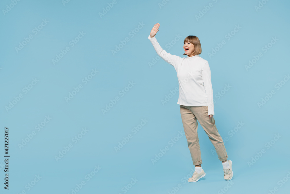 Full body side view elderly smiling cheerful caucasian woman 50s wear white knitted sweater walking going waving hand isolated on plain blue color background studio portrait. People lifestyle concept. Full body side view elderly smiling cheerful caucasian woman 50s wear white knitted sweater walking going waving hand isolated on plain blue color background studio portrait. People lifestyle concept.