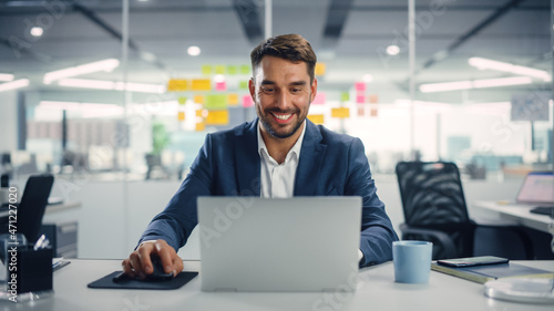 Young Happy Businessman Using Computer in Modern Office with Colleagues. Successful Handsome Manager Smiling, Working on Financial and Marketing Projects. Drinking Tea or Coffee from a Mug.