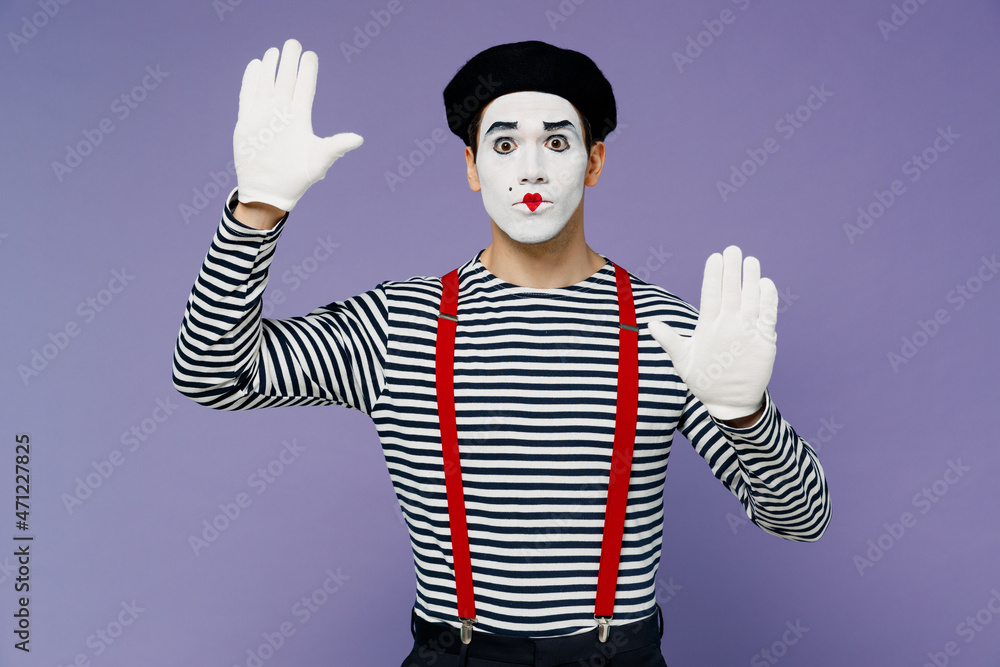 Bright young mime man with white face mask wears striped shirt beret looking camera hands raised as if touch stand near invisible wall isolated on plain pastel light violet background studio portrait.