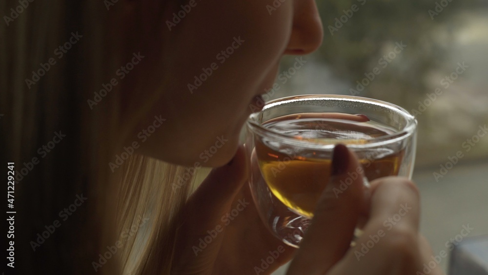 A young girl is sitting near the window and holding a glass mug with warm tea blowing cool air into her cup A young girl is sitting near the window and holding a glass mug with warm tea blowing cool air into her cup