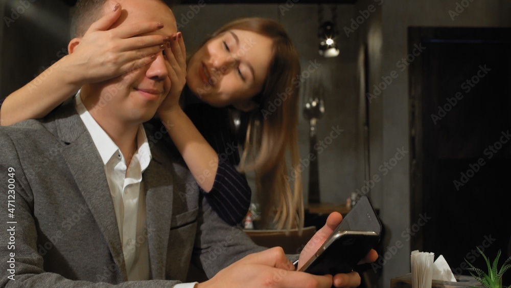 Fototapeta premium A man is sitting at a table in a cafe, browsing web pages on the Internet, a woman comes up from behind and covers his eyes with her hands and sits down at his table