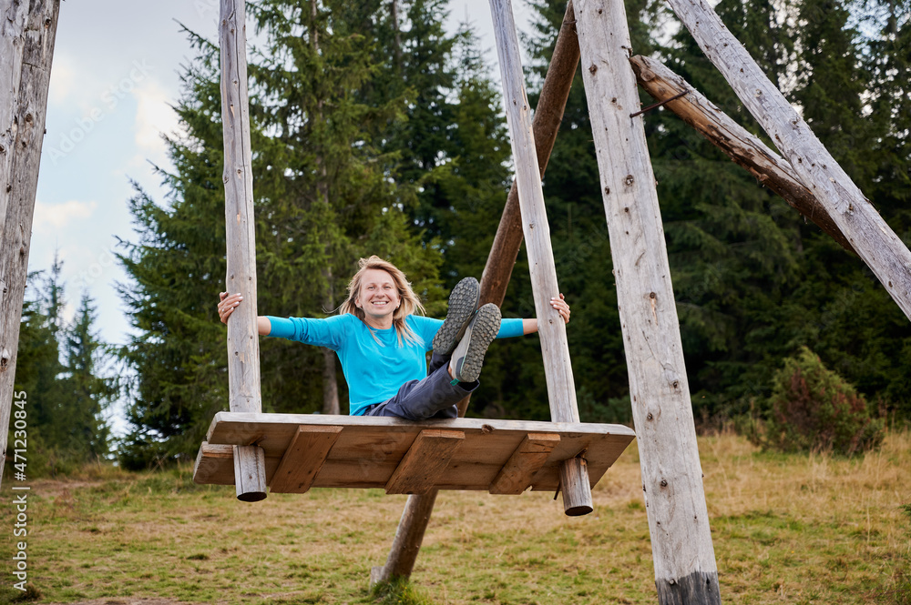 Happy woman sitting with straight legs on wooden seat attached to large wooden poles which she covers with her hands. Swinging on giant wooden swing on fresh air. Happy woman sitting with straight legs on wooden seat attached to large wooden poles which she covers with her hands. Swinging on giant wooden swing on fresh air.