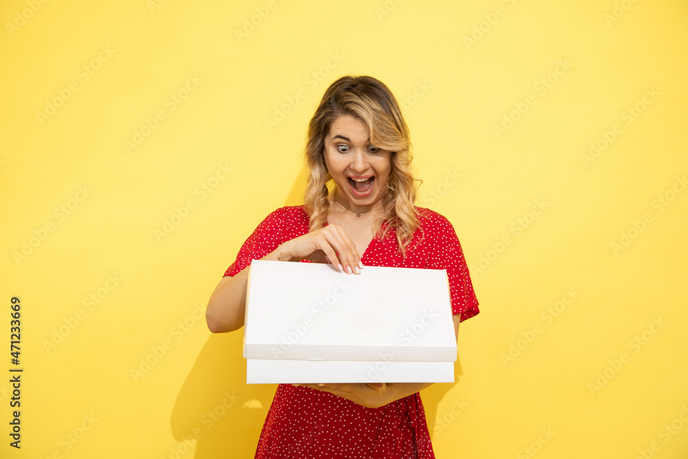 Young, beautiful long-haired blonde woman in red dress, joyfully opens white box with space for an inscription. Surprise concept. Love shopping, shopaholic. Young, beautiful long-haired blonde woman in red dress, joyfully opens white box with space for an inscription. Surprise concept. Love shopping, shopaholic.