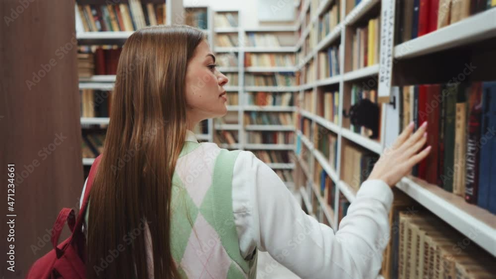Female student walking among rows of bookcases with backpack on shoulder, touching some of book covers. Following shot of woman in library. Concept of education Female student walking among rows of bookcases with backpack on shoulder, touching some of book covers. Following shot of woman in library. Concept of education