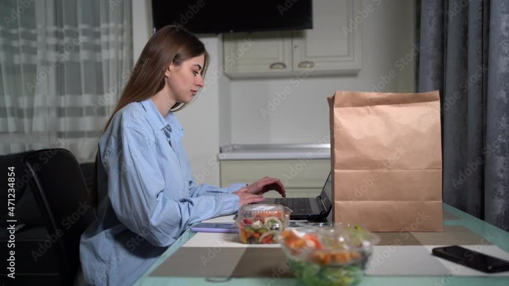 A Caucasian woman sits at home alone at a laptop in the evening in home clothes and works. food in disposable dishes is on the table near the paper bag. Food delivery concept. A Caucasian woman sits at home alone at a laptop in the evening in home clothes and works. food in disposable dishes is on the table near the paper bag. Food delivery concept.