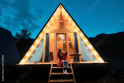 Leinwand Poster Woman drinking tea on the porch of a wooden lodge with lights of garlands in the evening
