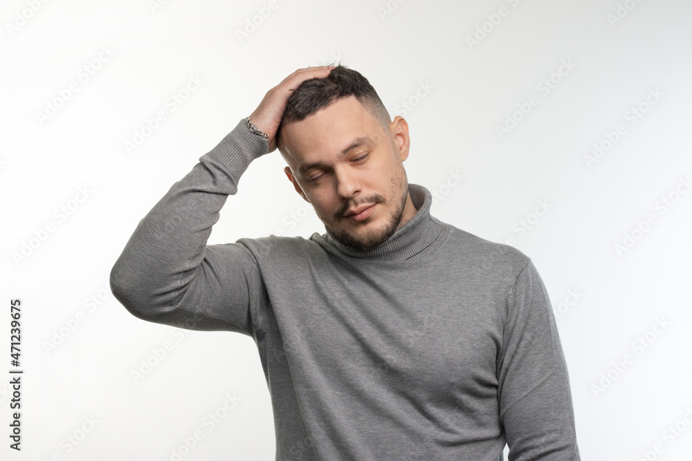 A young dark-haired man with a small beard holds his hand on his head, closed his eyes and thinks about something. The concept of nostalgia, memories. Light background. One person, isolated. A young dark-haired man with a small beard holds his hand on his head, closed his eyes and thinks about something. The concept of nostalgia, memories. Light background. One person, isolated.