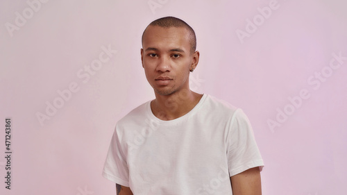 Portrait of young mixed race man in casual white t shirt looking at camera, standing isolated over pink background