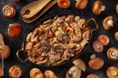 Beef stroganoff, mushroom and meat ragout in a creamy sauce, in a pan with ingredients, overhead flat lay shot on a dark background