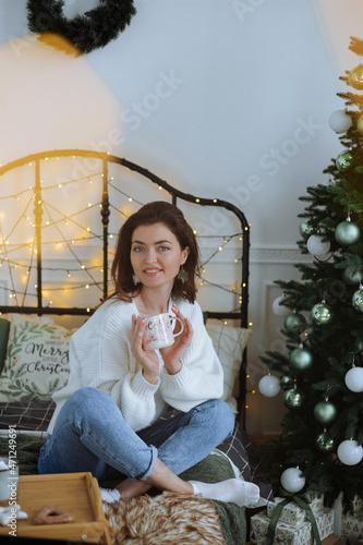 A beautiful girl is sitting on the bed, smiling, holding a mug. New Year and Christmas near the Christmas tree. New Year's gifts and happiness.