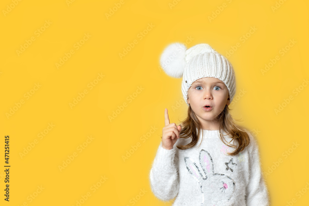 Portrait child girl with finger pointed up in winter knitted hat with pompon pose has idea isolated light yellow background. Christmas New Year shopping sale,promotion Portrait child girl with finger pointed up in winter knitted hat with pompon pose has idea isolated light yellow background. Christmas New Year shopping sale,promotion