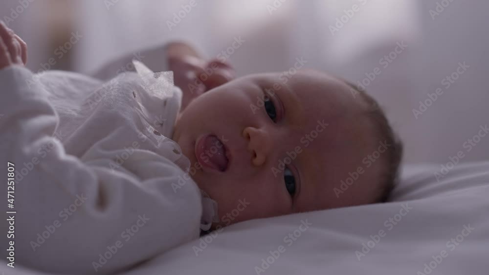 Close-up face little newborn baby girl lying on bed at home looking at camera showing tongue. Portrait of carefree Caucasian infant in white clothes resting indoors in bedroom. Slow motion Close-up face little newborn baby girl lying on bed at home looking at camera showing tongue. Portrait of carefree Caucasian infant in white clothes resting indoors in bedroom. Slow motion