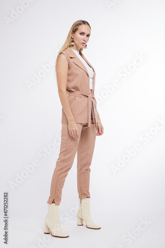 High fashion photo of a beautiful elegant young woman in a pretty beige suit, pants, trousers, vest, boots posing over white, soft gray background. Studio Shot, portrait. Blonde