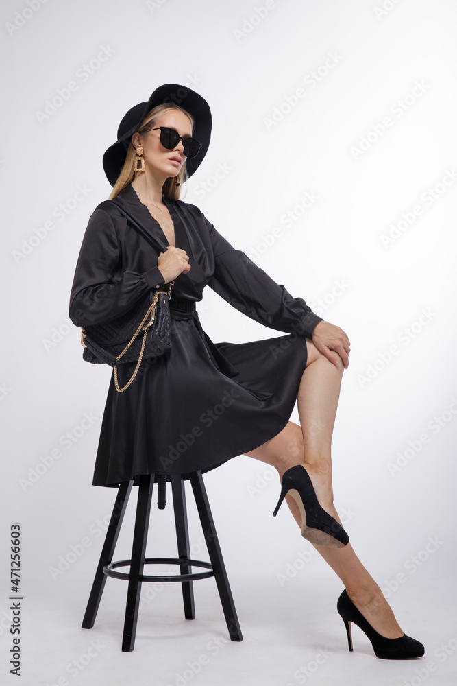 High fashion photo of a beautiful elegant young woman in a pretty black dress, hat, heels, sunglasses, bag posing over white, soft gray background. Studio Shot, portrait. Model is sitting on a chair High fashion photo of a beautiful elegant young woman in a pretty black dress, hat, heels, sunglasses, bag posing over white, soft gray background. Studio Shot, portrait. Model is sitting on a chair