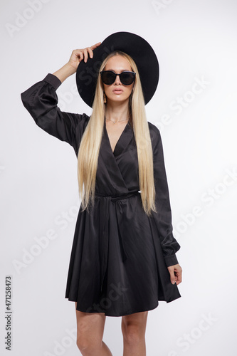 High fashion photo of a beautiful elegant young woman in a pretty black dress, hat, high heels, sunglasses, posing over white, soft gray background. Studio Shot, portrait. Blonde, slim figure