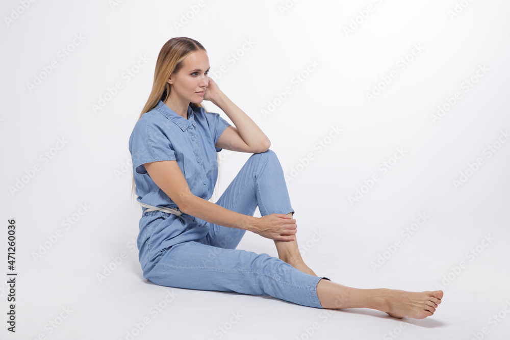 High fashion photo of a beautiful elegant young woman in a pretty blue jumpsuit, barefoot posing over white, soft gray background. Studio Shot, portrait. The model is sitting in the background. Blonde High fashion photo of a beautiful elegant young woman in a pretty blue jumpsuit, barefoot posing over white, soft gray background. Studio Shot, portrait. The model is sitting in the background. Blonde