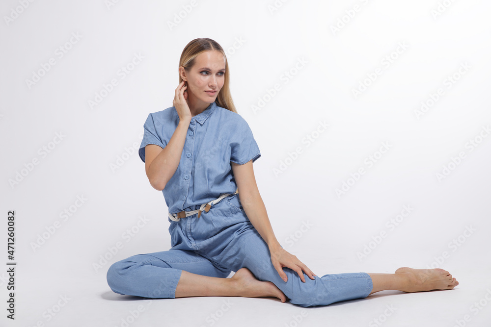 High fashion photo of a beautiful elegant young woman in a pretty blue jumpsuit, barefoot posing over white, soft gray background. Studio Shot, portrait. The model is sitting in the background. Blonde High fashion photo of a beautiful elegant young woman in a pretty blue jumpsuit, barefoot posing over white, soft gray background. Studio Shot, portrait. The model is sitting in the background. Blonde