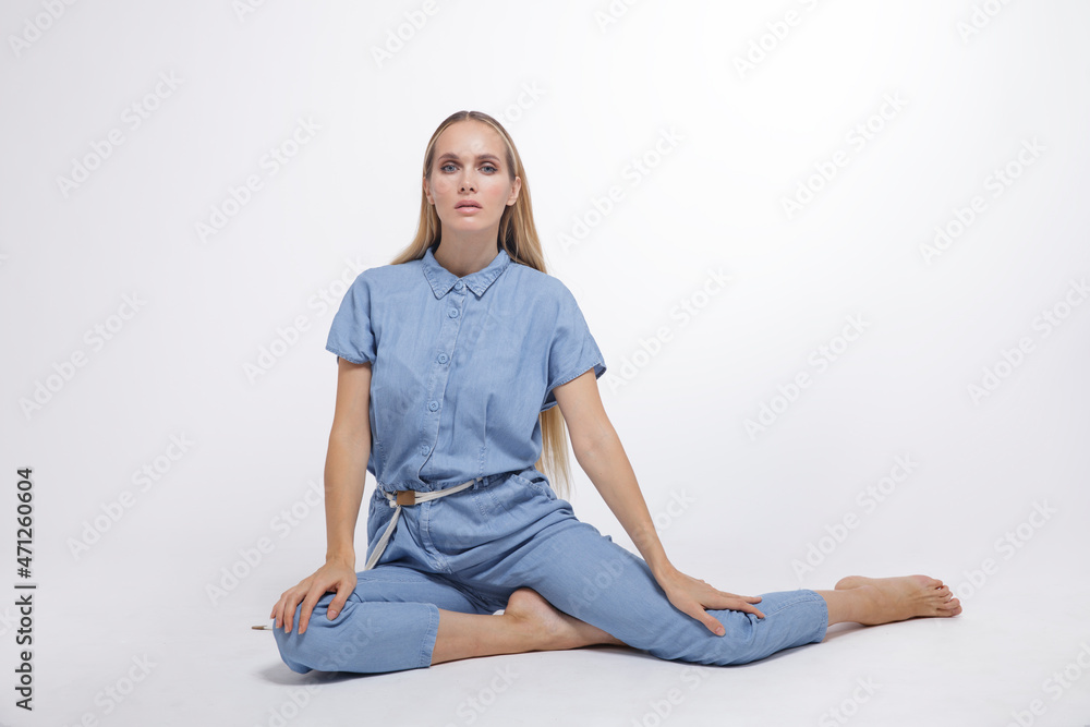 High fashion photo of a beautiful elegant young woman in a pretty blue jumpsuit, barefoot posing over white, soft gray background. Studio Shot, portrait. The model is sitting in the background. Blonde