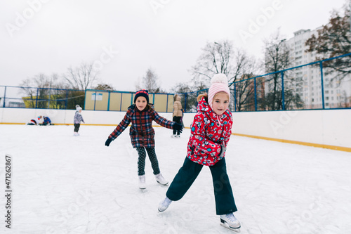 Little caucasian girls ice skating on rink in city in wintertime, moving, smiling, spreading arms, laughing. Full lengh horizontal shot. Happy childhood and winter activities concept