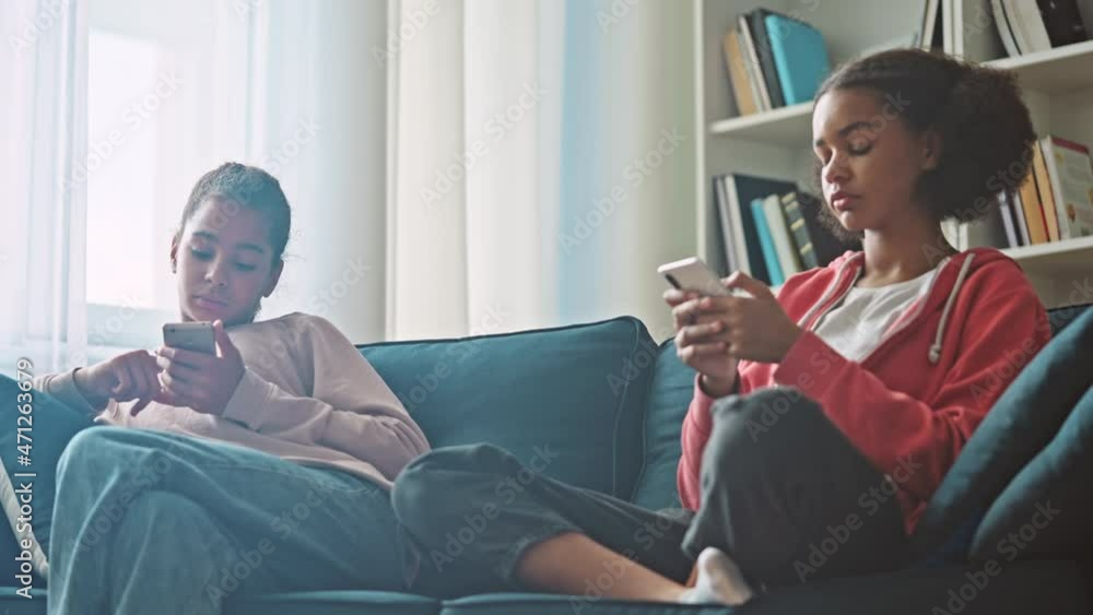 Two teenage girls sitting on couch with smartphones, browsing internet at home Two teenage girls sitting on couch with smartphones, browsing internet at home