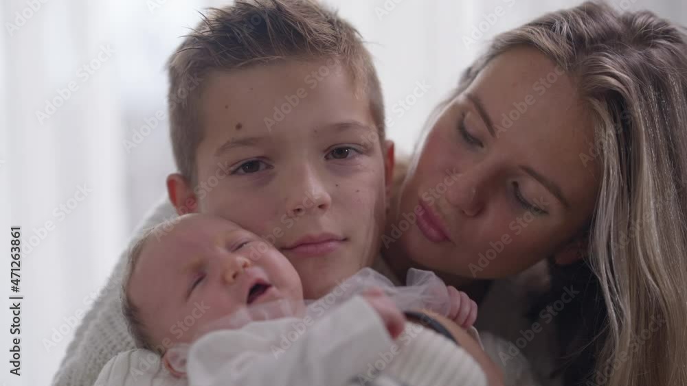 Teenage boy looking at camera standing indoors with crying baby sister and beautiful mother. Portrait of Caucasian brother calming down infant with woman at home posing. Slow motion Teenage boy looking at camera standing indoors with crying baby sister and beautiful mother. Portrait of Caucasian brother calming down infant with woman at home posing. Slow motion