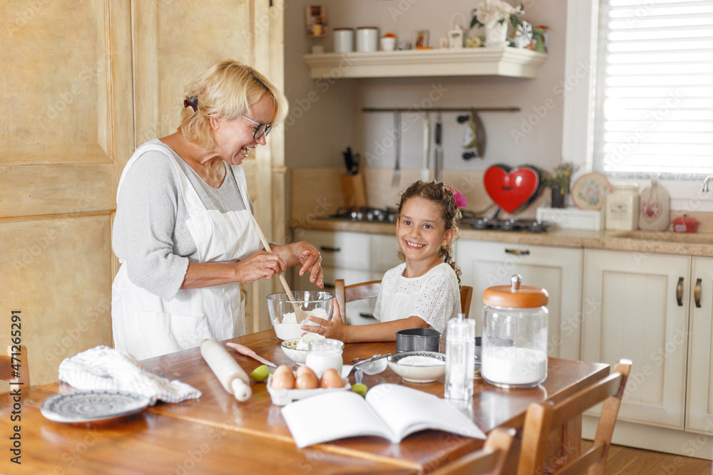 Grandmother and pretty adorable granddaughter prepare together in the kitchen, with ingredients on the table. Grandmother and pretty adorable granddaughter prepare together in the kitchen, with ingredients on the table.