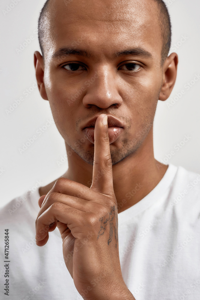 Portrait of dark skinned young guy showing shh gesture, keeping a secret, posing isolated over white background