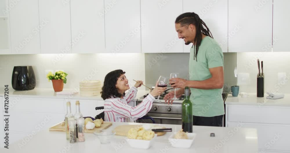 Happy biracial woman in wheelchair preparing food and drinking wine with male partner in kitchen Happy biracial woman in wheelchair preparing food and drinking wine with male partner in kitchen