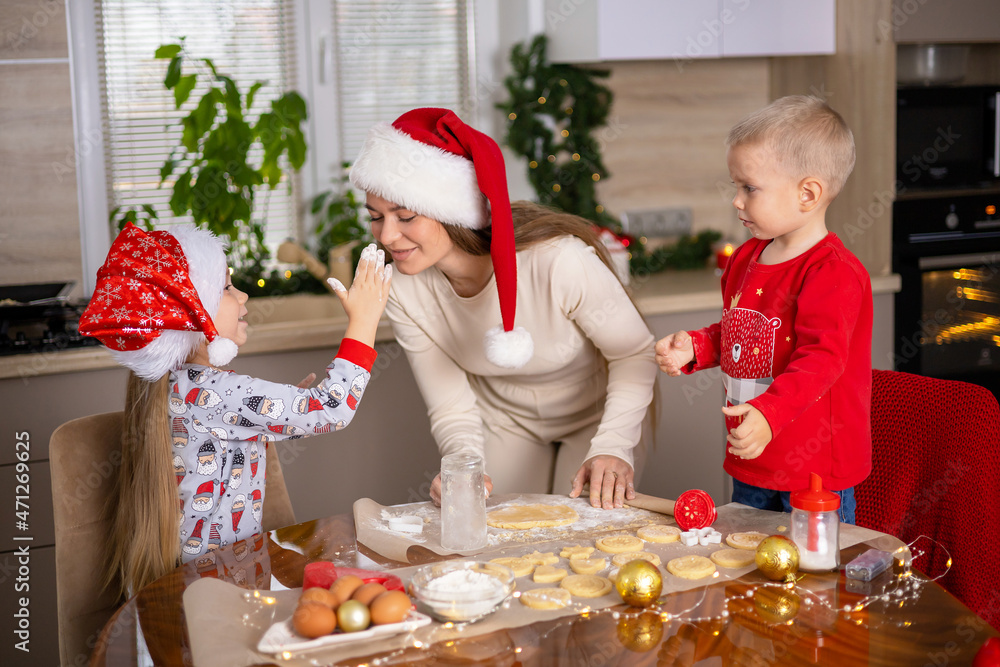 Merry Christmas. Happy Mom with children son and daughter bake Christmas cookies and stain each other with flour and laugh. Kitchen at home. Festive atmosphere