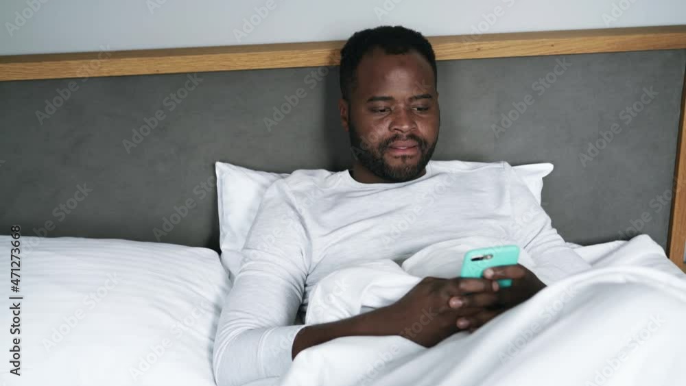 Pensive African man texting by phone and yawning in bed Pensive African man texting by phone and yawning in bed