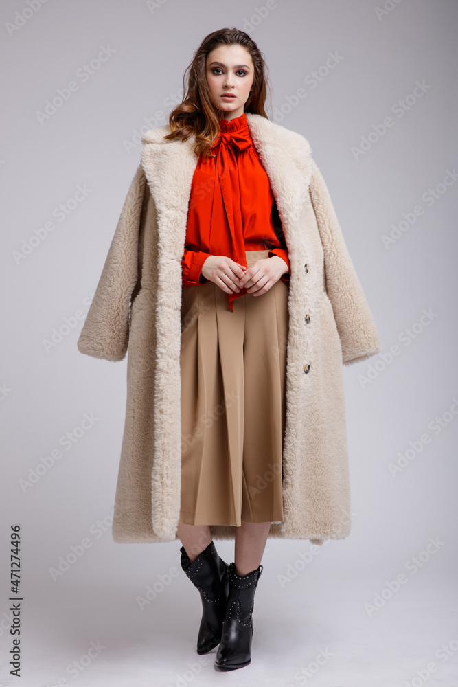 High fashion photo of a beautiful elegant young woman in a pretty beige coat, long skirt, red bow blouse, boots posing over white, soft gray background. Brown hair. Studio Shot, portrait. High fashion photo of a beautiful elegant young woman in a pretty beige coat, long skirt, red bow blouse, boots posing over white, soft gray background. Brown hair. Studio Shot, portrait.