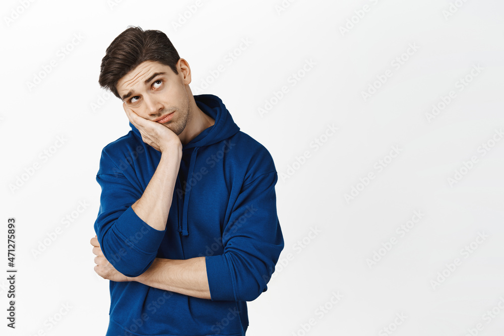 Bored or sad guy peeking at ceiling, man looking up with boredom face, standing over white background in blue hoodie Bored or sad guy peeking at ceiling, man looking up with boredom face, standing over white background in blue hoodie