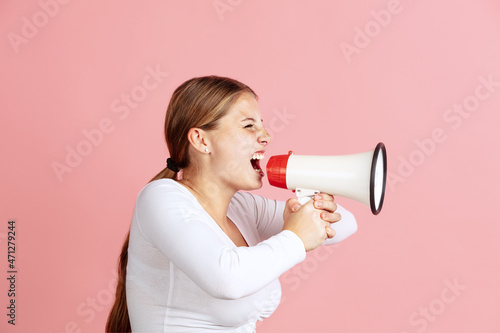 Portrait of young red-headed girl shouting at megaphone isolated over pink studio background. Profile view