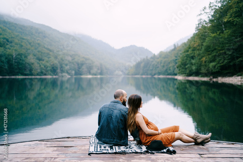 Man with pregnant woman are sitting on a pier by the lake in the park