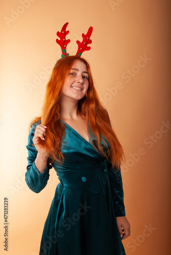 a red-haired girl on an orange background stands in isolation in a Christmas decor with reindeer horns for the new year and Christmas 2022 2021