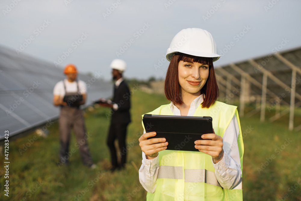 Caucasian woman in helmet using digital tablet for work on solar station. African american businessman and indian technician talking behind. Green energy concept.