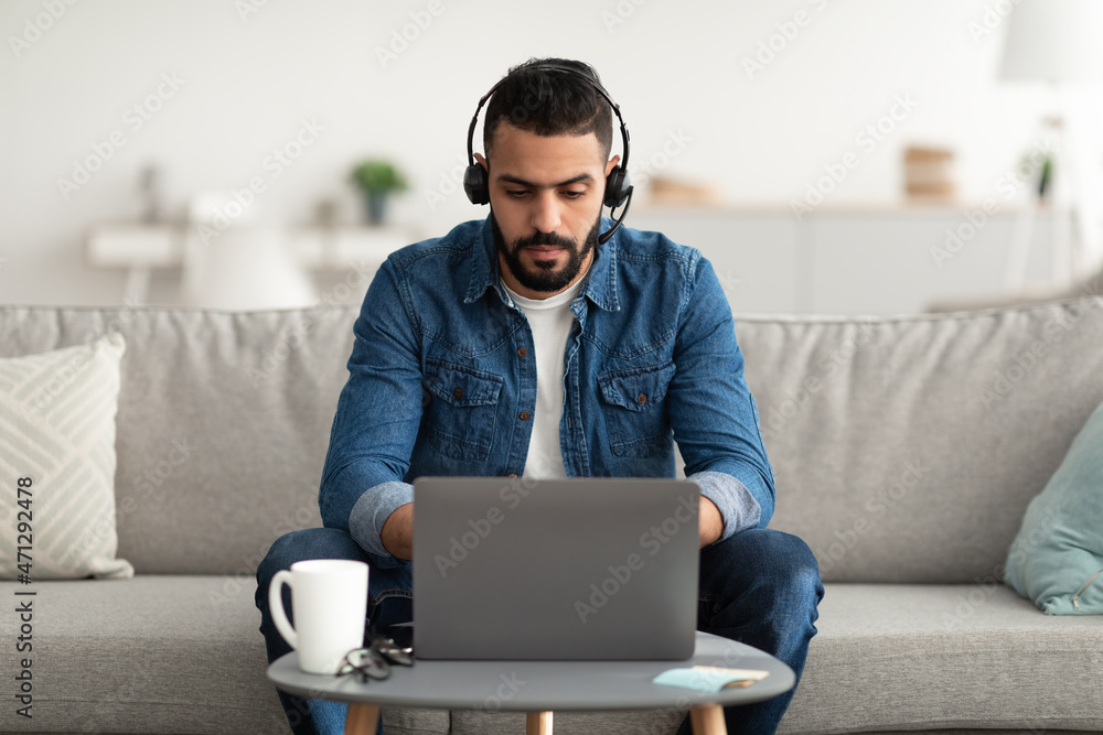 Young serious Arab man wearing headphones, using laptop computer, working online from home Young serious Arab man wearing headphones, using laptop computer, working online from home