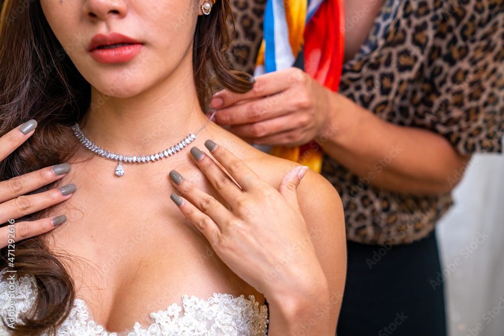 Asian LGBTQ guy make-up artist wearing diamond necklace to female bride in wedding dress in fitting room at wedding studio. Small business entrepreneur bridal shop owner and wedding planner concept