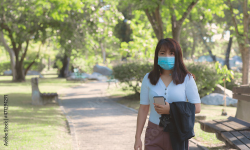 attractive business woman wearing face mask hold smartphone standing in park with sunrise and blur nature background. business new normal with corona virus or covid-19.