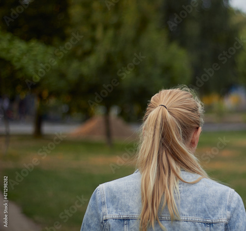 Rear view of a young charming blonde in a denim jacket. Nature and park background.
