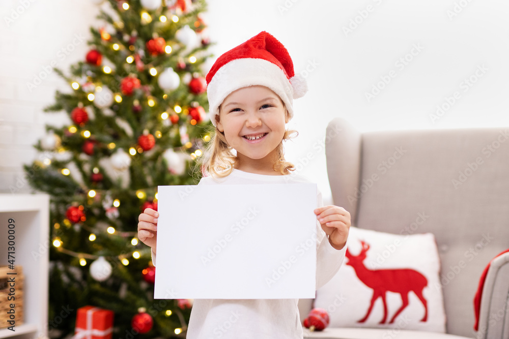 Little girl holding blank paper near Christmas tree at home Little girl holding blank paper near Christmas tree at home